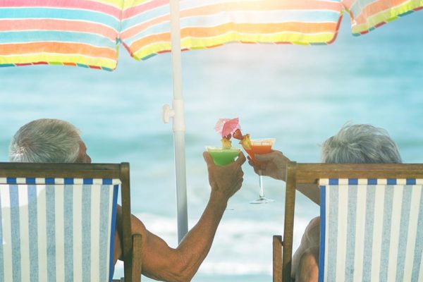 Seniorenpaar sitzt in Klappstühlen am Strand unter einem bunten Sonnenschirm und genießt Cocktails | Wohnen im Alter