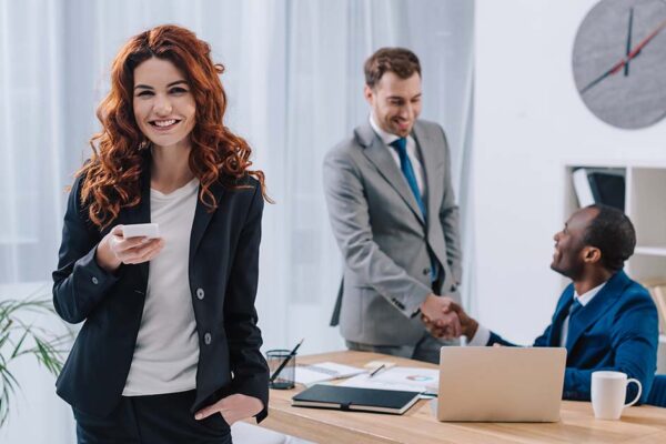 Eine junge Frau steht in einem Büro und ist glücklich über einen neuen Job, im Hintergrund schütteln sich zwei Männer die Hand | Immobilienverkauf bei Jobwechsel