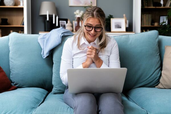 Eine junge Frau sitzt mit dem Laptop auf ihren Beinen auf dem Sofa in ihrer Mietwohnung und freut sich, eine Immobilie zum Kauf gefunden zu haben, die sie sich leisten kann | Immobilie kaufen